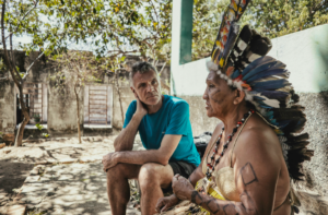 Homem de camiseta azul e bermuda preta, com expressão atenta, escuta uma liderança indígena que está sentada ao seu lado. A liderança veste um cocar colorido feito de penas, colares de miçangas e tem pinturas corporais nos braços e no rosto. Eles estão sentados em uma área externa, com chão de terra, árvores e uma construção simples ao fundo, sob luz natural.