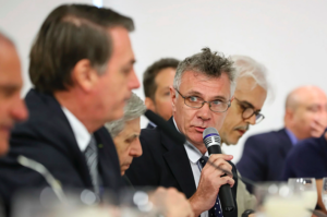 Em uma reunião formal, um homem de terno preto, camisa branca e gravata azul com bolinhas segura um microfone e fala, olhando atentamente para alguém fora do quadro. Ele está cercado por outros homens também de terno, entre eles o ex-presidente Jair Bolsonaro, que aparece desfocado em primeiro plano, olhando na direção do orador. A mesa está posta com copos e microfones, indicando um evento oficial ou institucional.