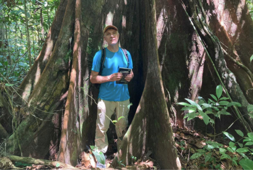 Dom Phillips em pé na floresta amazônica, com boné, camiseta azul e mochila, segurando um caderno, posicionado entre as raízes de uma grande árvore.