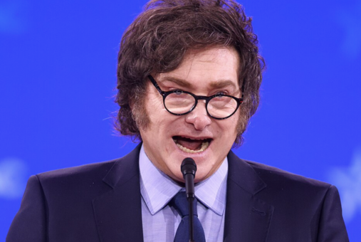 Argentine President Javier Milei speaking passionately at a podium, wearing glasses, a dark suit, and a blue tie, with a blue background behind him.