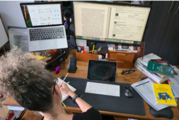 Brazilian journalist Cecília Olliveira seen from behind at her home office desk, working on a notepad while surrounded by monitors, documents, and books related to organized crime and public security.