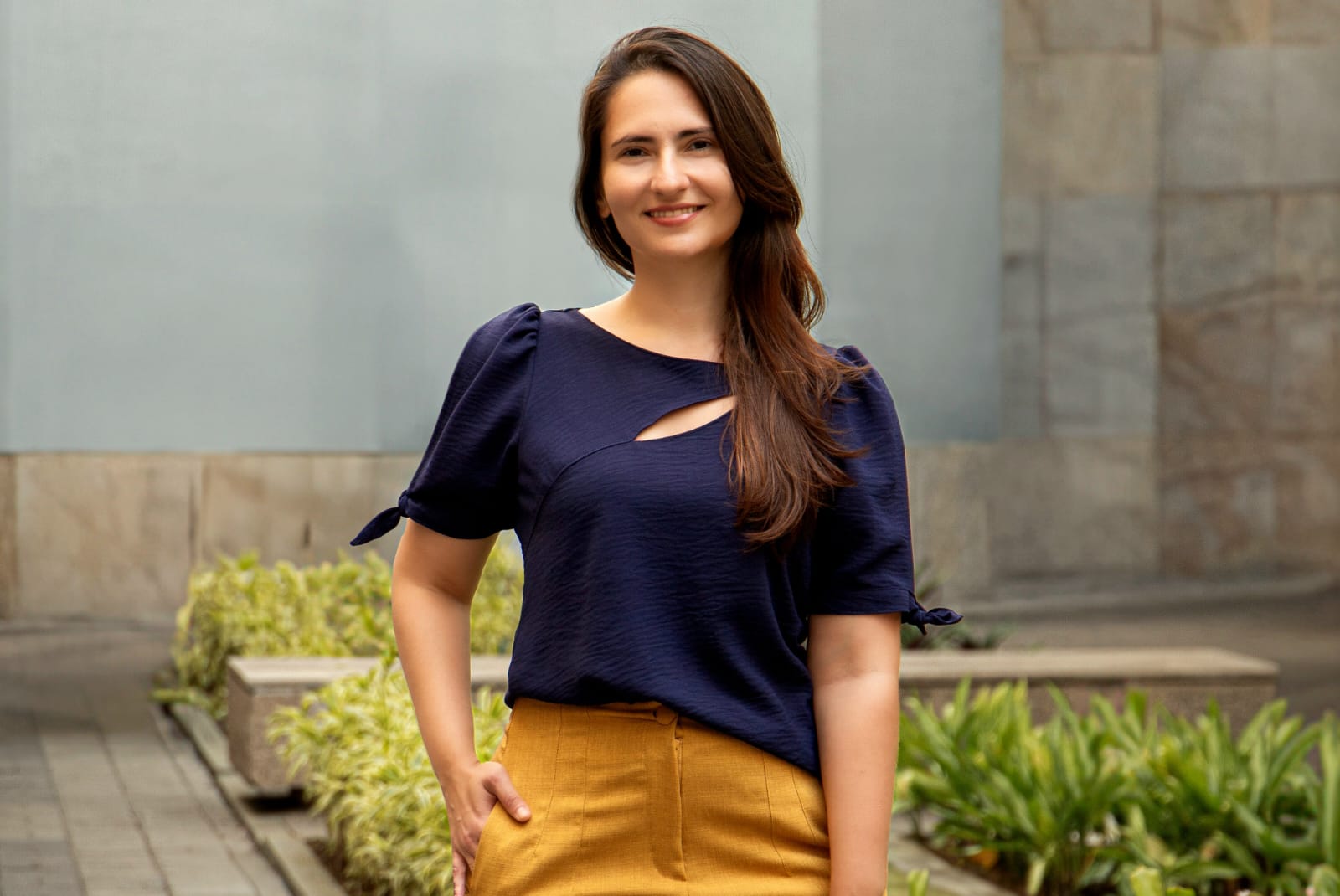 Laren Aniceto, a young woman with long brown hair, stands smiling with one hand in her pocket, wearing a navy blouse and mustard pants