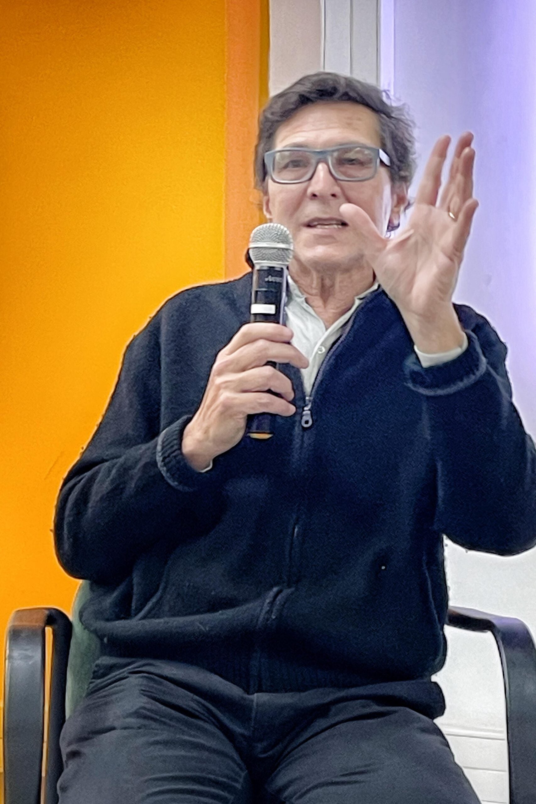Fernando Ruiz speaks into a microphone while gesturing with his hand during a public event. He is seated, wearing glasses and a dark jacket, against a background divided into bright orange and light purple.