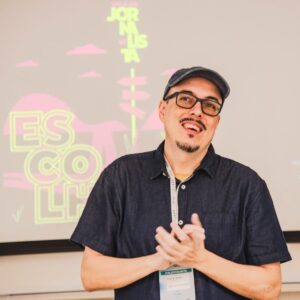 Hombre de mediana edad con gorra negra y camisa negra frente a un tablero sonriendo y aplaudiendo