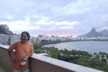 Marcelo Beraba smiles, leaning against the railing of his apartment overlooking Rodrigo de Freitas Lagoon and the Dois Irmãos mountain in Rio de Janeiro.