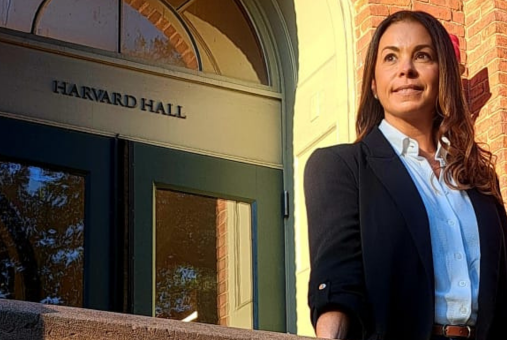 Journalist Simone Iglesias standing in front of Harvard Hall, wearing a dark blazer and white shirt, with sunlight casting a warm glow.