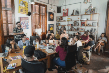 A group of people gathered around a large table in a brightly lit room, with bookshelves and posters on the walls, participating in a newsroom meeting.
