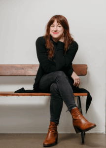 Woman sitting on a wooden bench with legs crossed, wearing a black outfit and brown boots, smiling at the camera.