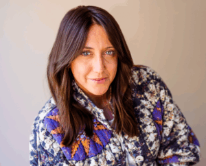 Woman with long brown hair looking at the camera, wearing a patterned jacket and sitting against a neutral background.
