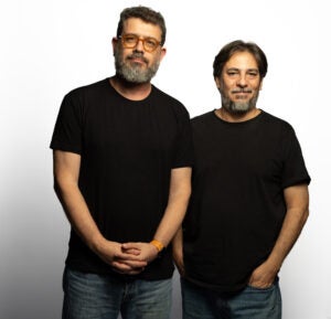 Two men standing side by side against a plain background, both wearing black shirts and looking at the camera.