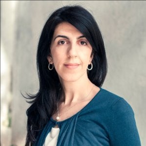 Woman with long dark hair, wearing a blue top and hoop earrings, poses in front of a softly lit neutral background.