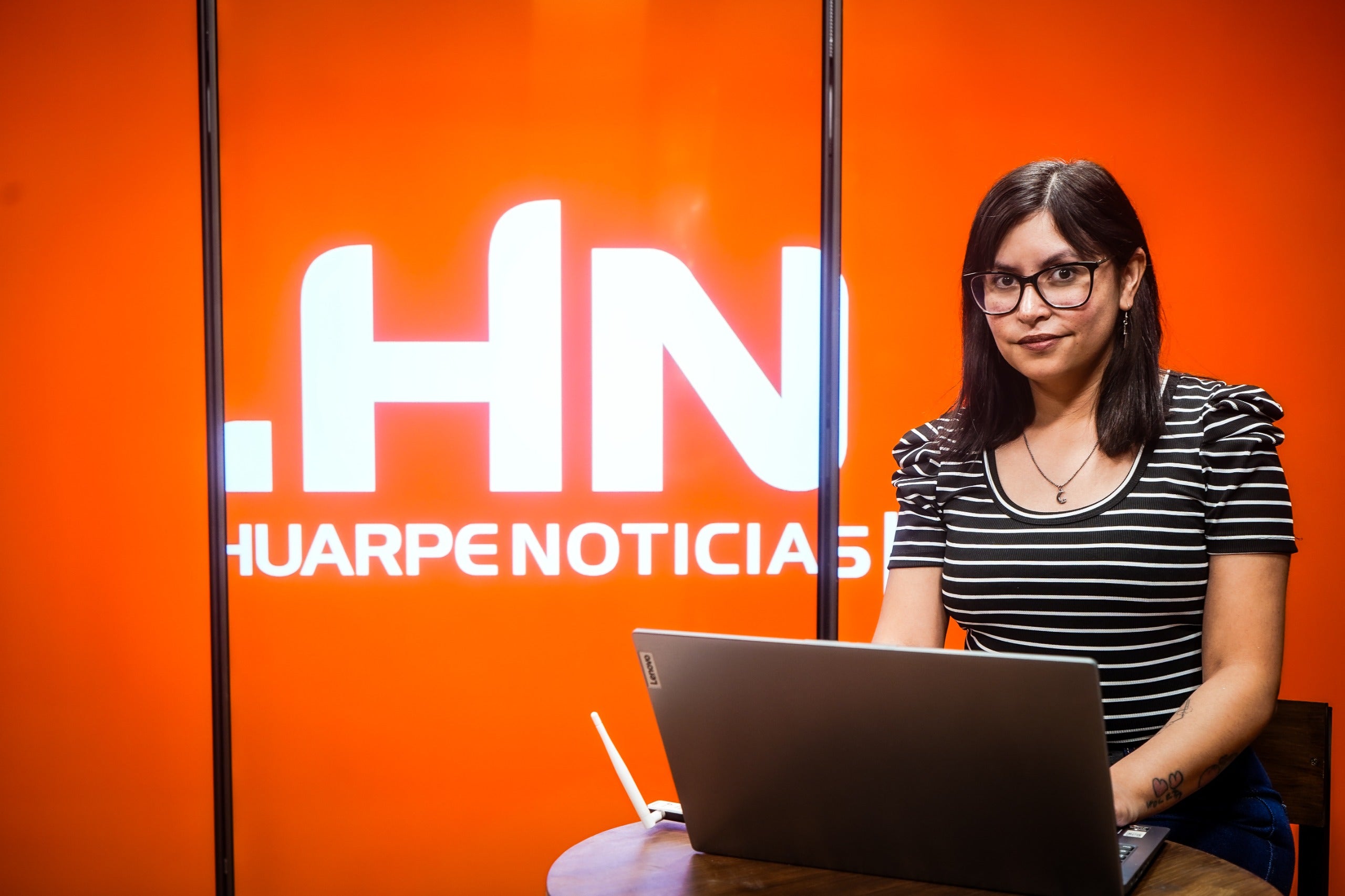 Argentine journalist Yanina Paez poses with a laptop in front of an orange background displaying the logo of the Huarpe Noticias media outlet.