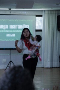 Woman with a baby on her hip talks at front of room