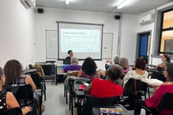 A classroom scene in Niterói, Brazil, shows journalism educator Victor Terra leading a media literacy session for older adults. Participants, mostly women over 60, sit facing a projection screen that reads “Let’s think together?” as they discuss internet use and information habits.