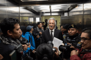 Man in black suit and tie stands amid reporters in elevator