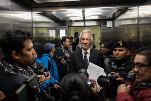 Man in black suit and tie stands amid reporters in elevator