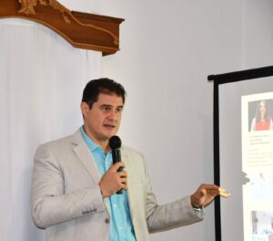 Hombre de traje beige con camisa azul hablando a una audiencia con un micrófono en su mano