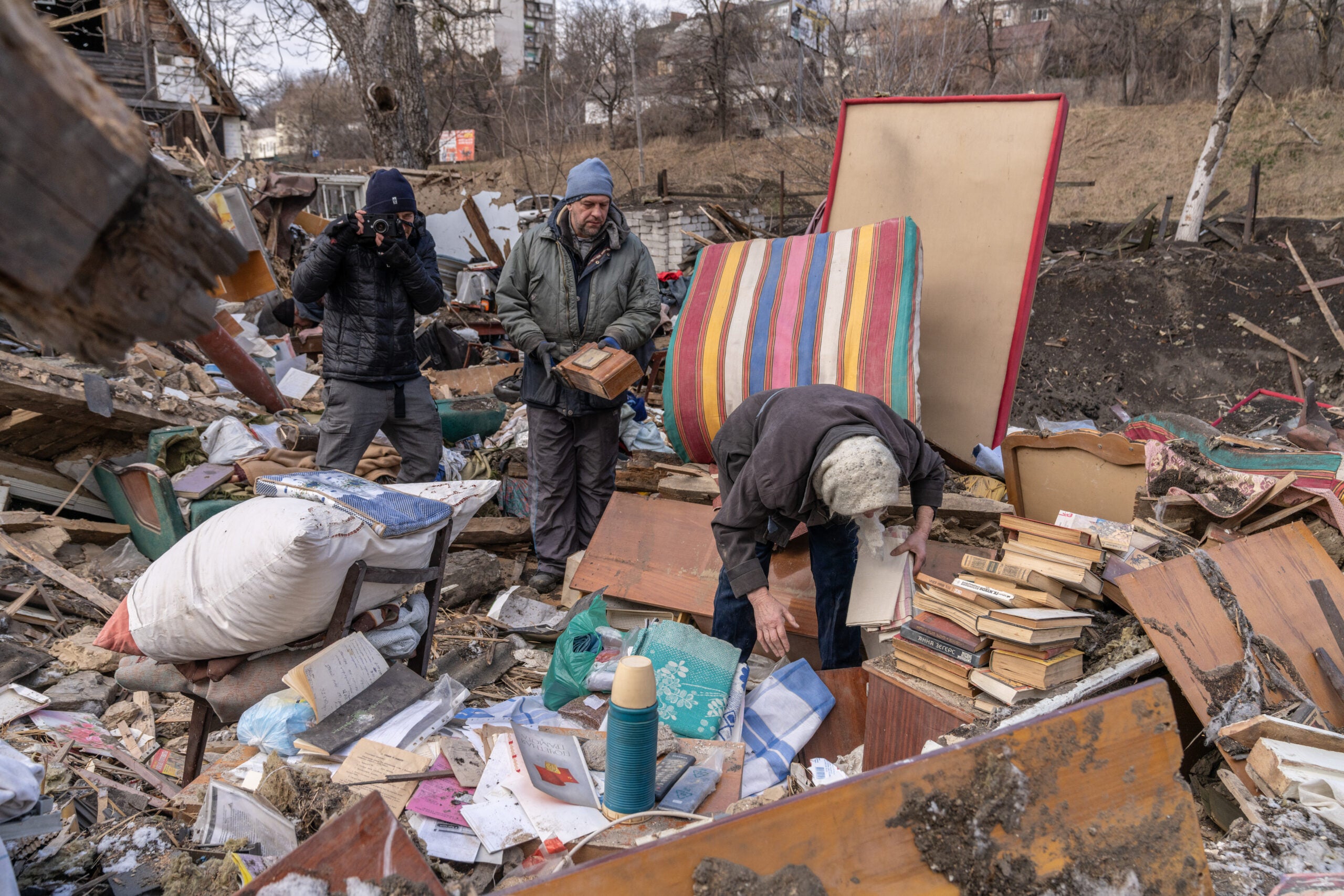 Two people looking for objects in ruins behind them a man with a camera