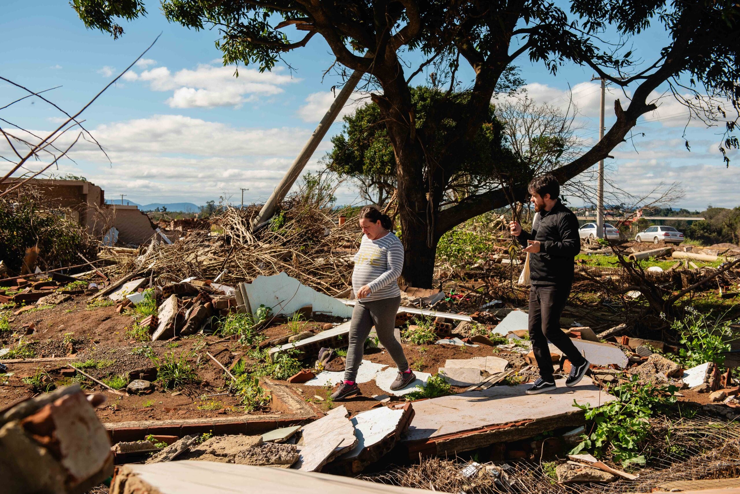 Man and woman walk through destroyed houses and trees